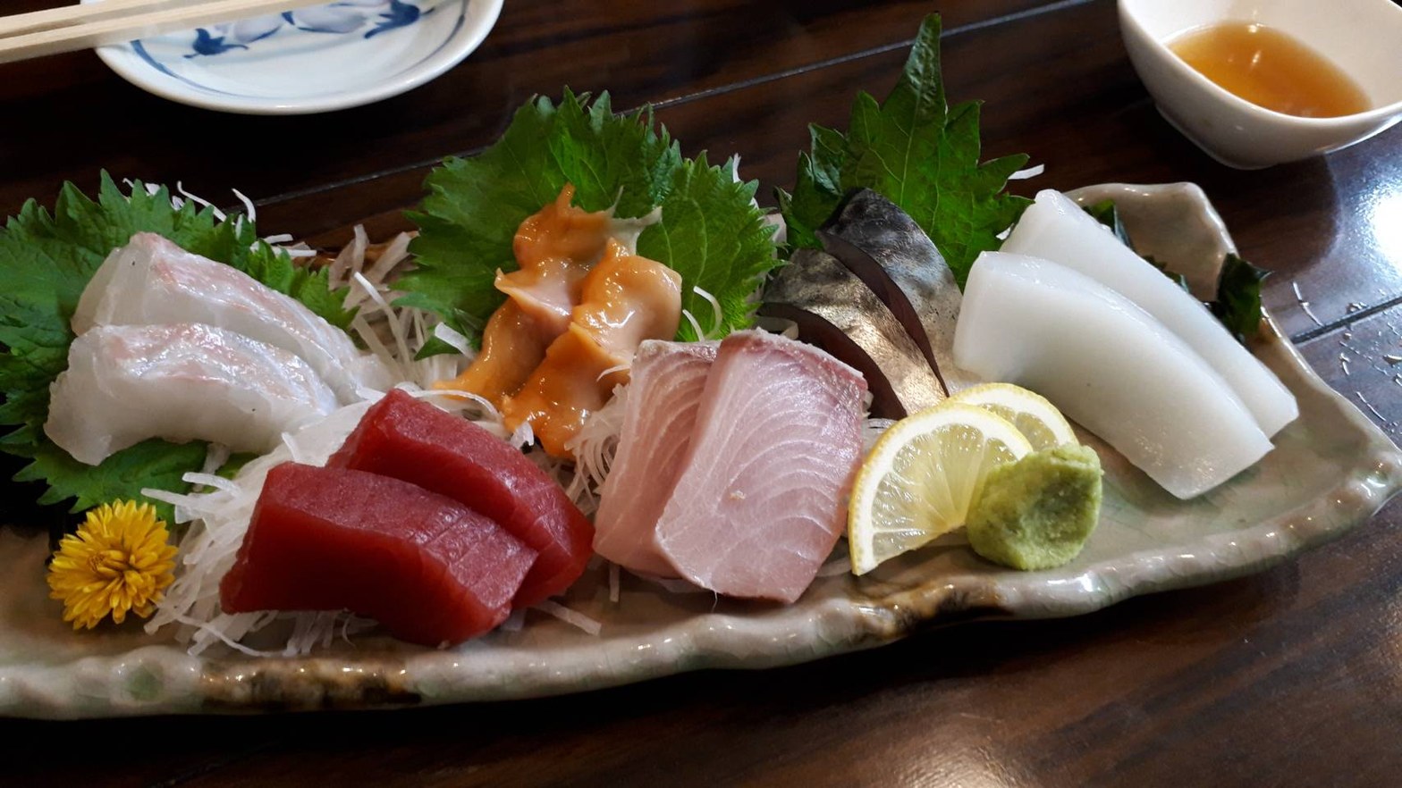 白い長皿に並ぶ刺し盛り。鰤・鮪・鯵・鯖・烏賊・貝類と黄菊の花、レモン、わさびが盛られている