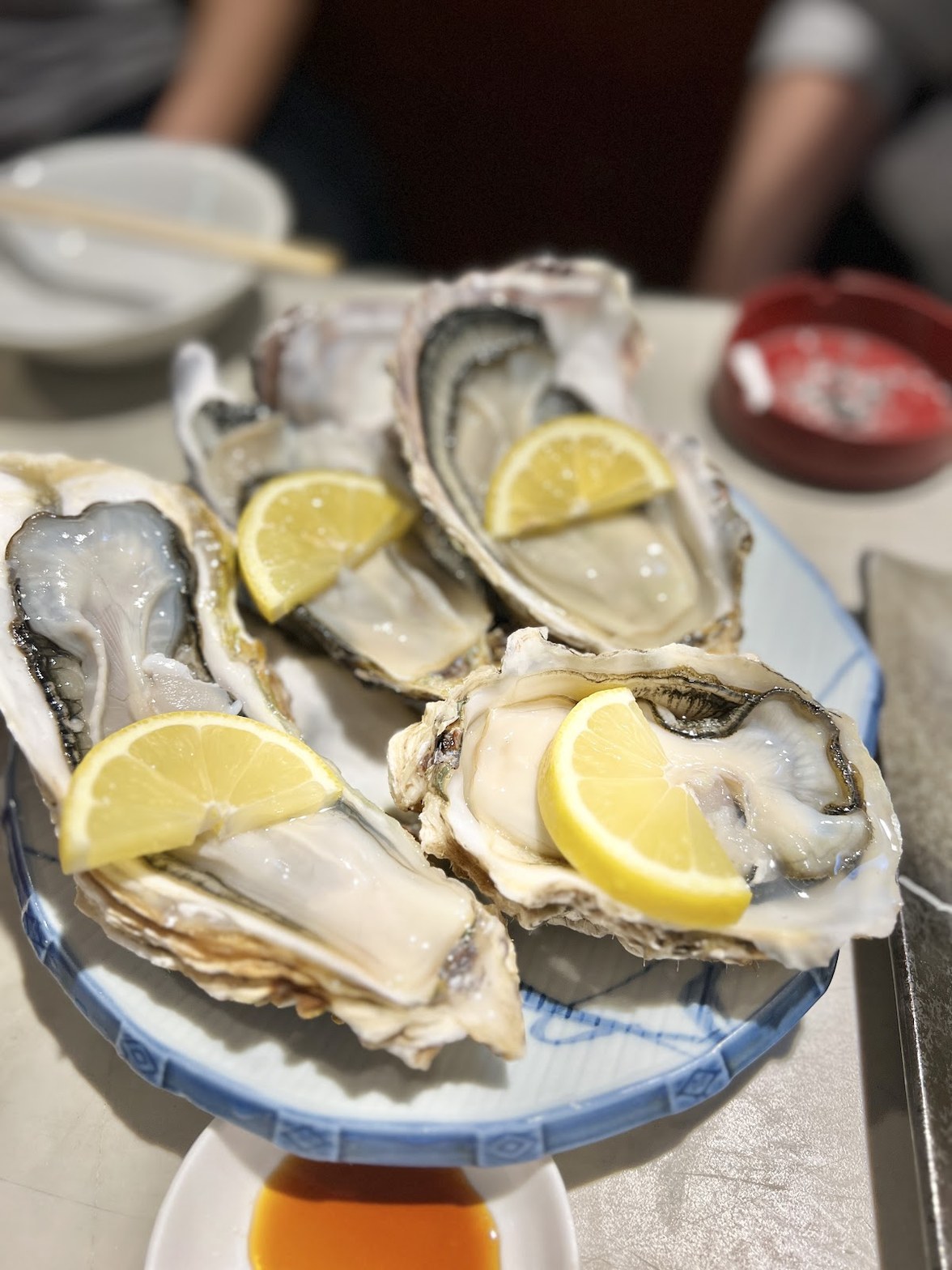 丸い白皿に並ぶ牡蠣の生食4個。殻付きのまま黄色いレモンのくし切りを添えて盛られている